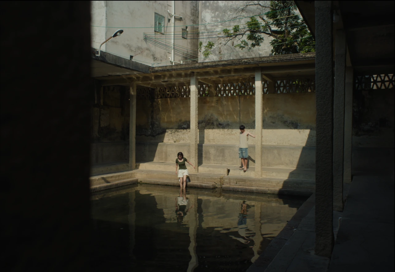 Two figures beside a reflecting pool in a courtyard, framed by a shaded passageway.