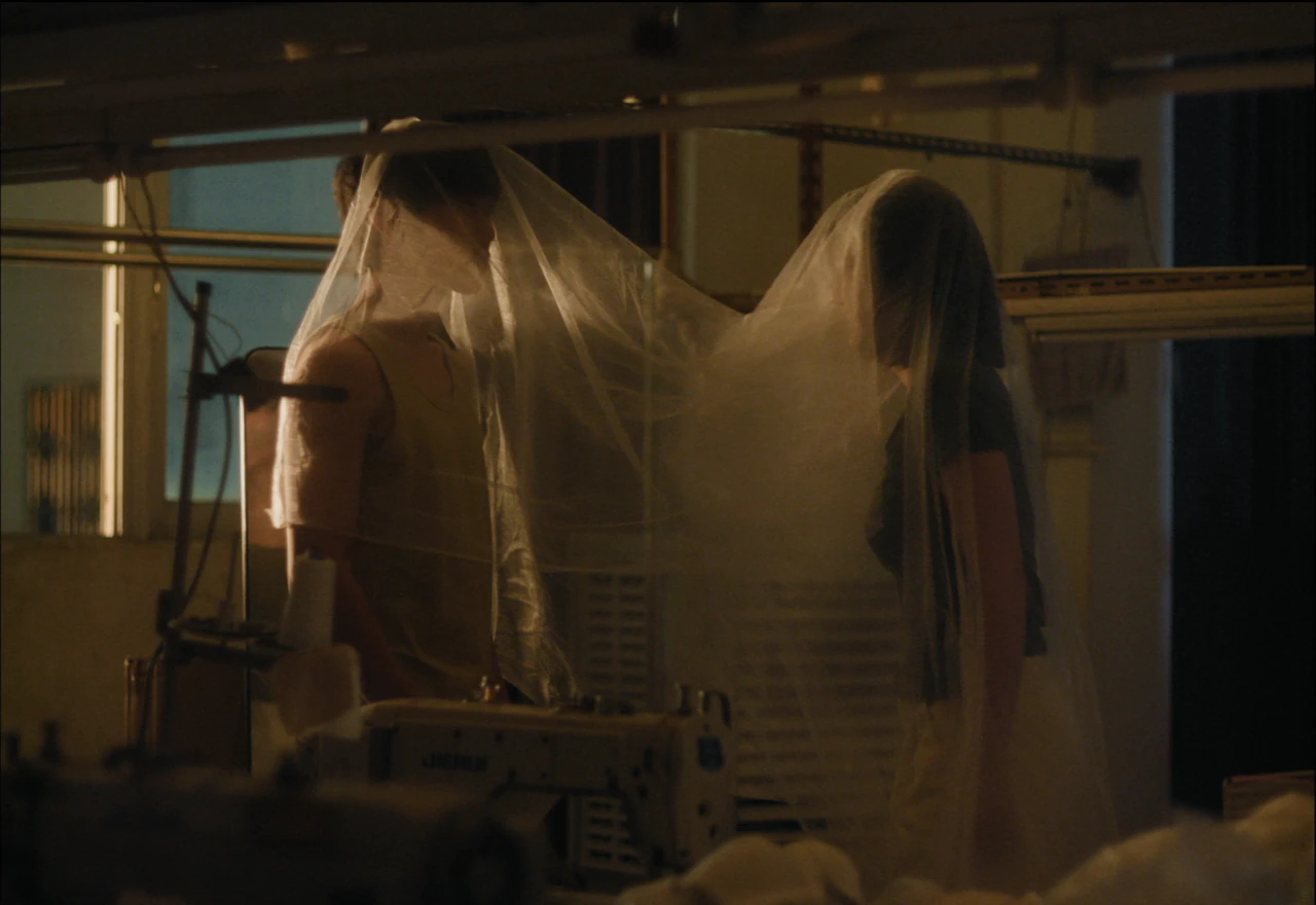 Two figures draped in sheer fabric between the machines of a garment factory.