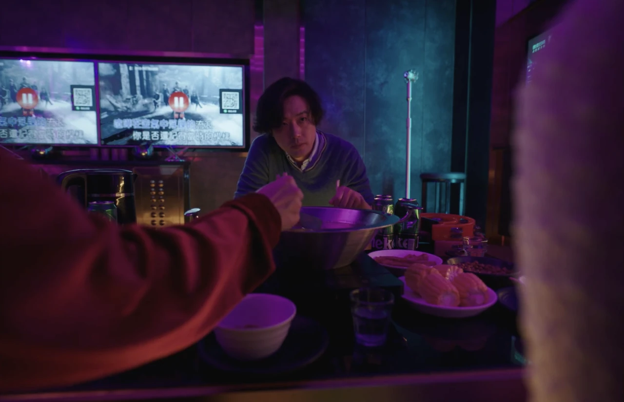 A man sits across a table in a purple-lit room as someone reaches toward him.