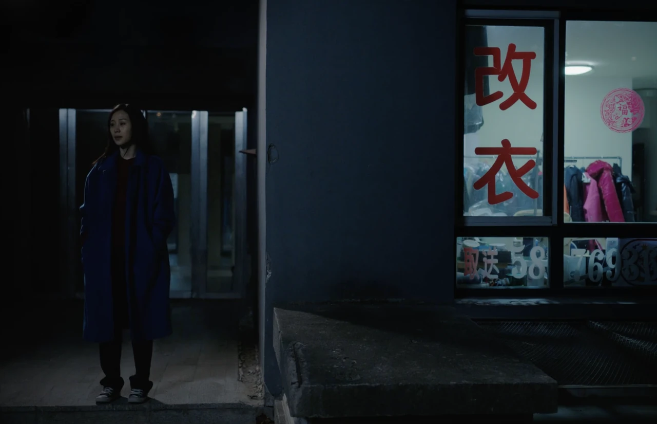 A woman in a blue coat stands outside a tailoring shop at night.